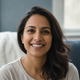 Headshot of a smiling South Asian woman in her late 30s, conveying relief and well-being, with a blurred background of soft b